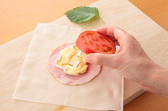 カマンベールチーズは4等分にする。トマトはへたを取り、4等分の輪切りにする。春巻きの皮の中央にイタリアンパセリの葉を1枚おき、上にハム1枚をのせる。カマンベールチーズ1/4量を平らにのせ、トマト1枚、青じそ1枚を順に重ねる。ハムとトマトではさむようにカマンベールチーズを重ねると、揚げ焼きしたときにはみ出しにくくなる。また、イタリアンパセリが透けて見えるよう、最初におくのもポイント。