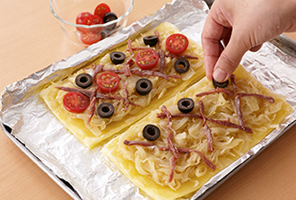 1のパイシートの上に玉ねぎを敷き、アンチョビを格子状になるように並べる。すき間にオリーブ、プチトマトを彩りよく置く。オーブントースターで5分ほど、こんがりするまで焼く。