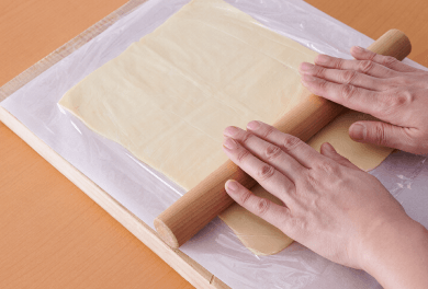 まな板を縦に置き、オーブン用シートを敷いて、半解凍させた冷凍パイシートをのせ、上にラップをかける。綿棒でのばして長さ25～28センチくらいに伸ばしてラップを外す。